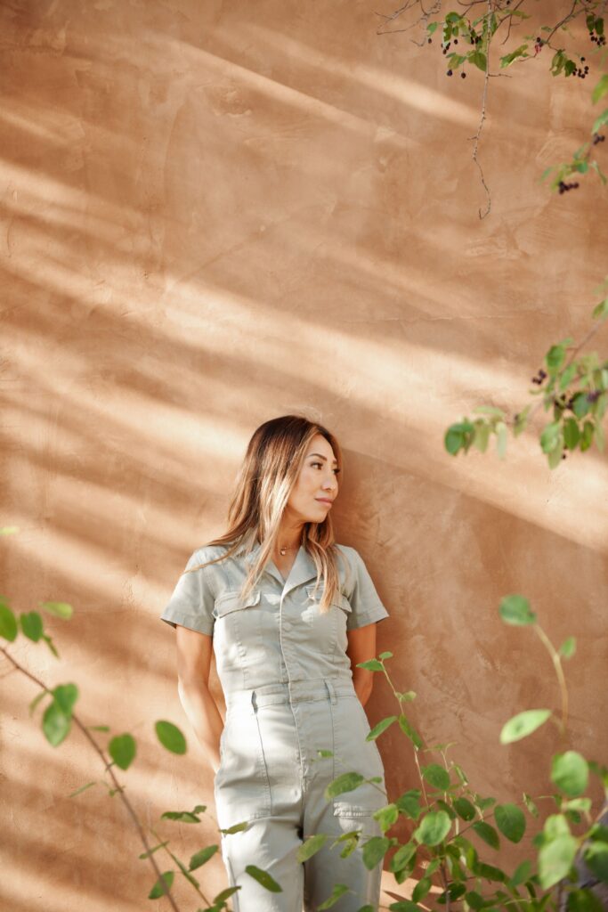 Woman in a jumpsuit leaning against an adobe wall.