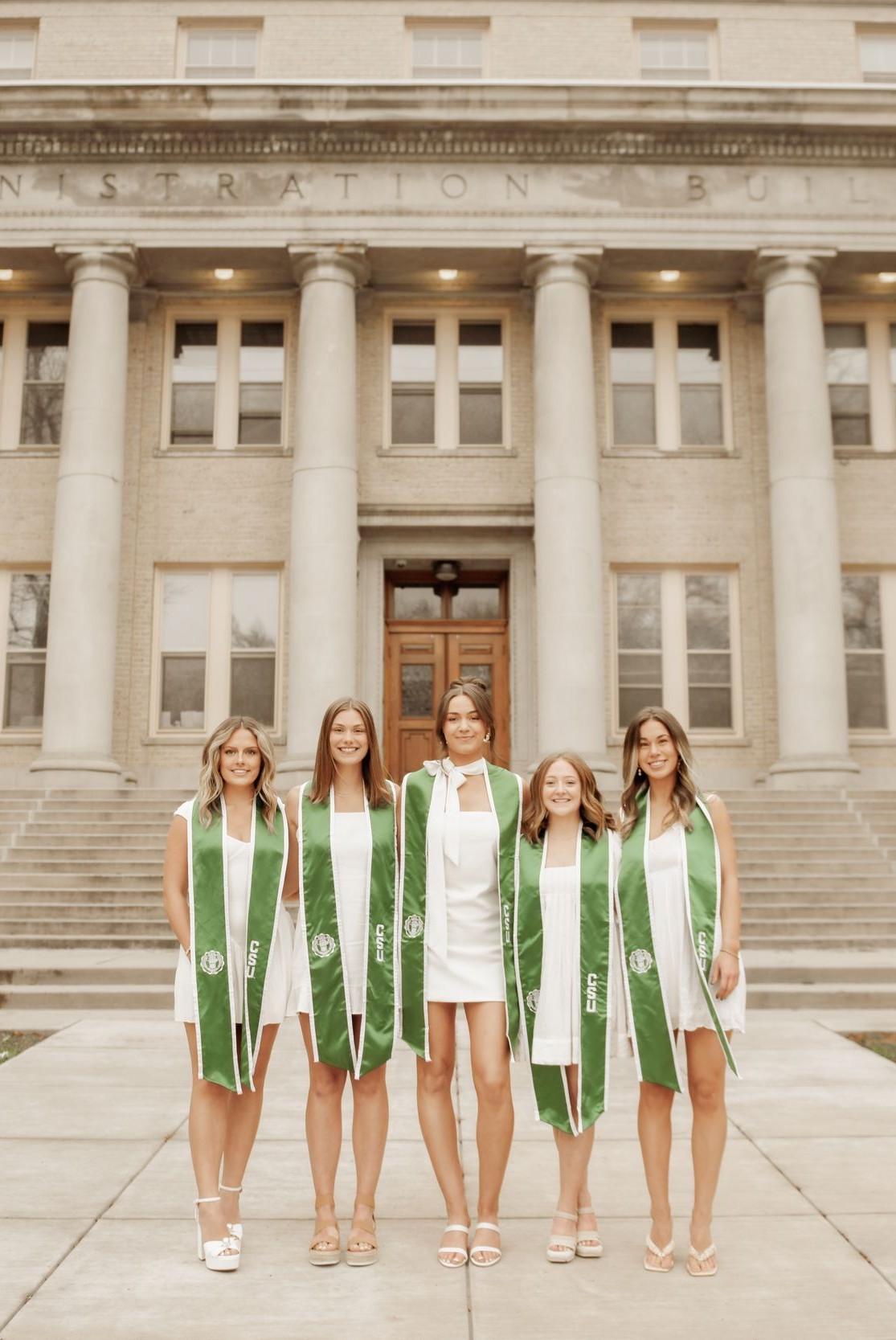 Five graduates in dresses and CSU stoles standing in front of the campus Administration Building.