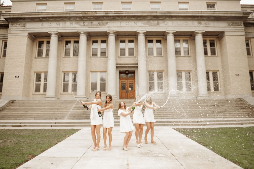 Five women in white dresses spraying champagne in front of a columned administration building.