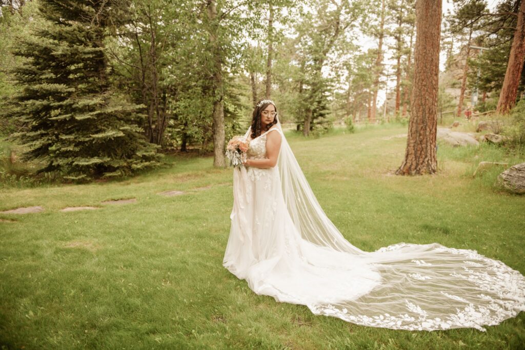 Bride in a lace wedding gown holding a bouquet with a long flowing veil in a forest setting.