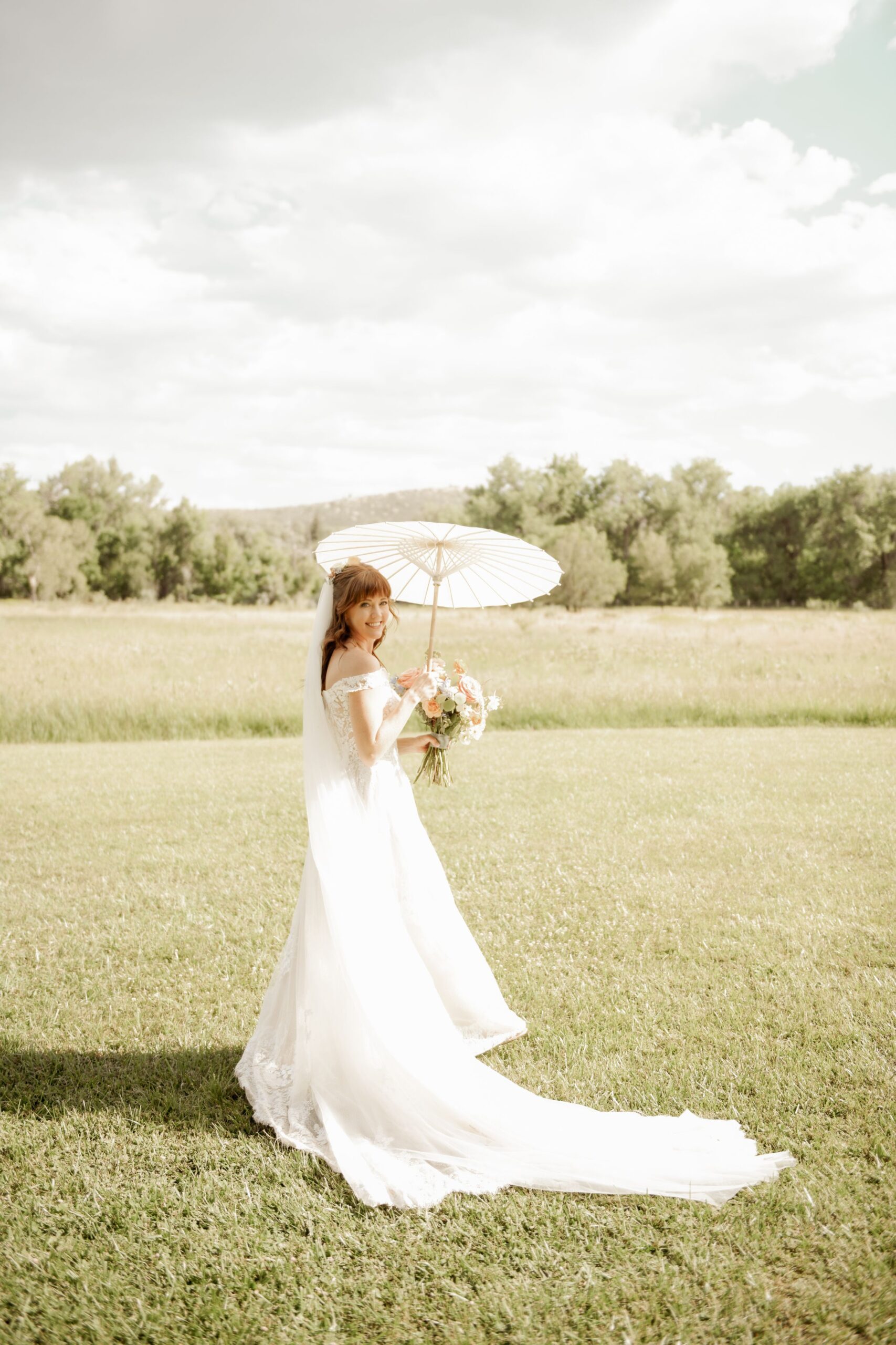 Bride in an off-shoulder gown holding a parasol and bouquet in an open field