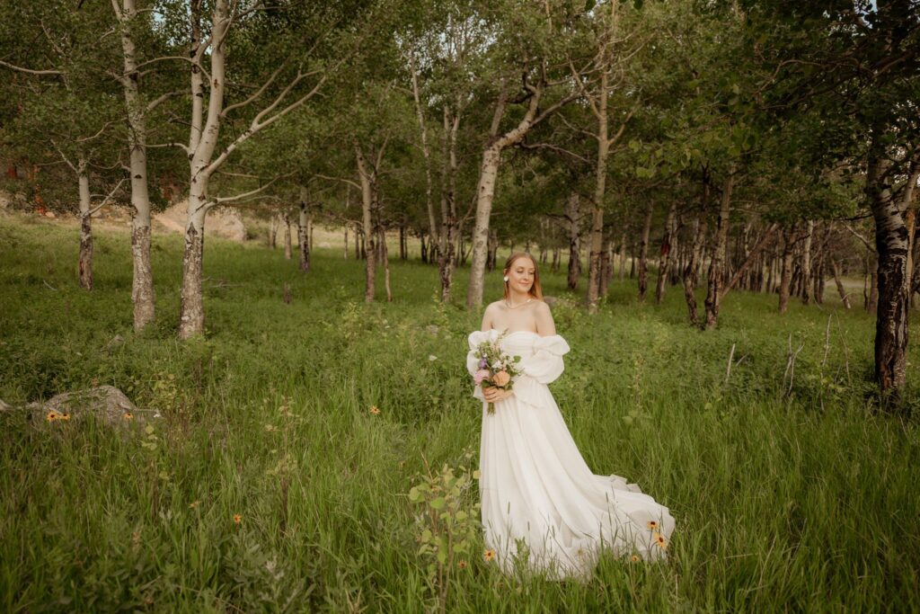 Bride in an off-shoulder wedding gown standing in a forest clearing holding a bouquet
