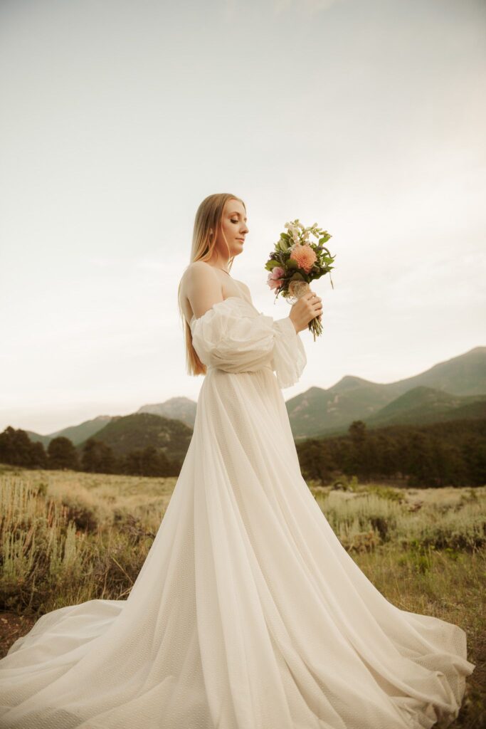 Bride holding a bouquet in a flowing wedding dress with mountain views behind her.