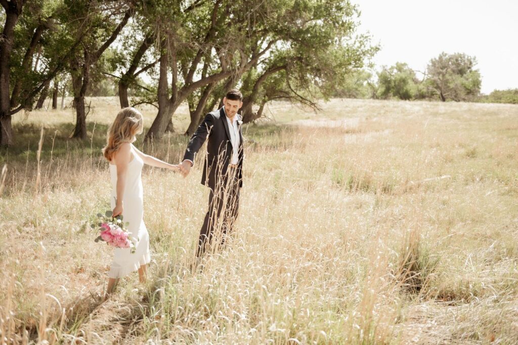 Bride and groom walking hand in hand through a sunlit field.