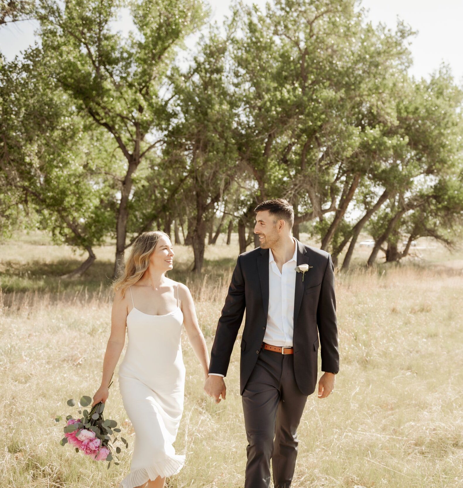 Bride and groom walking hand in hand through a sunlit meadow with a bouquet.