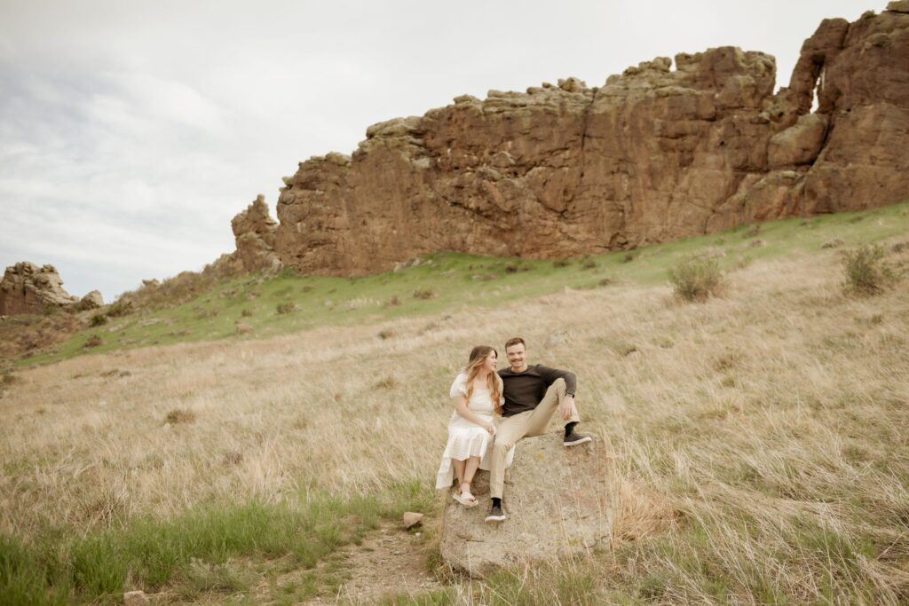 Couple sitting on a rock in a grassy field near rugged cliffs.
