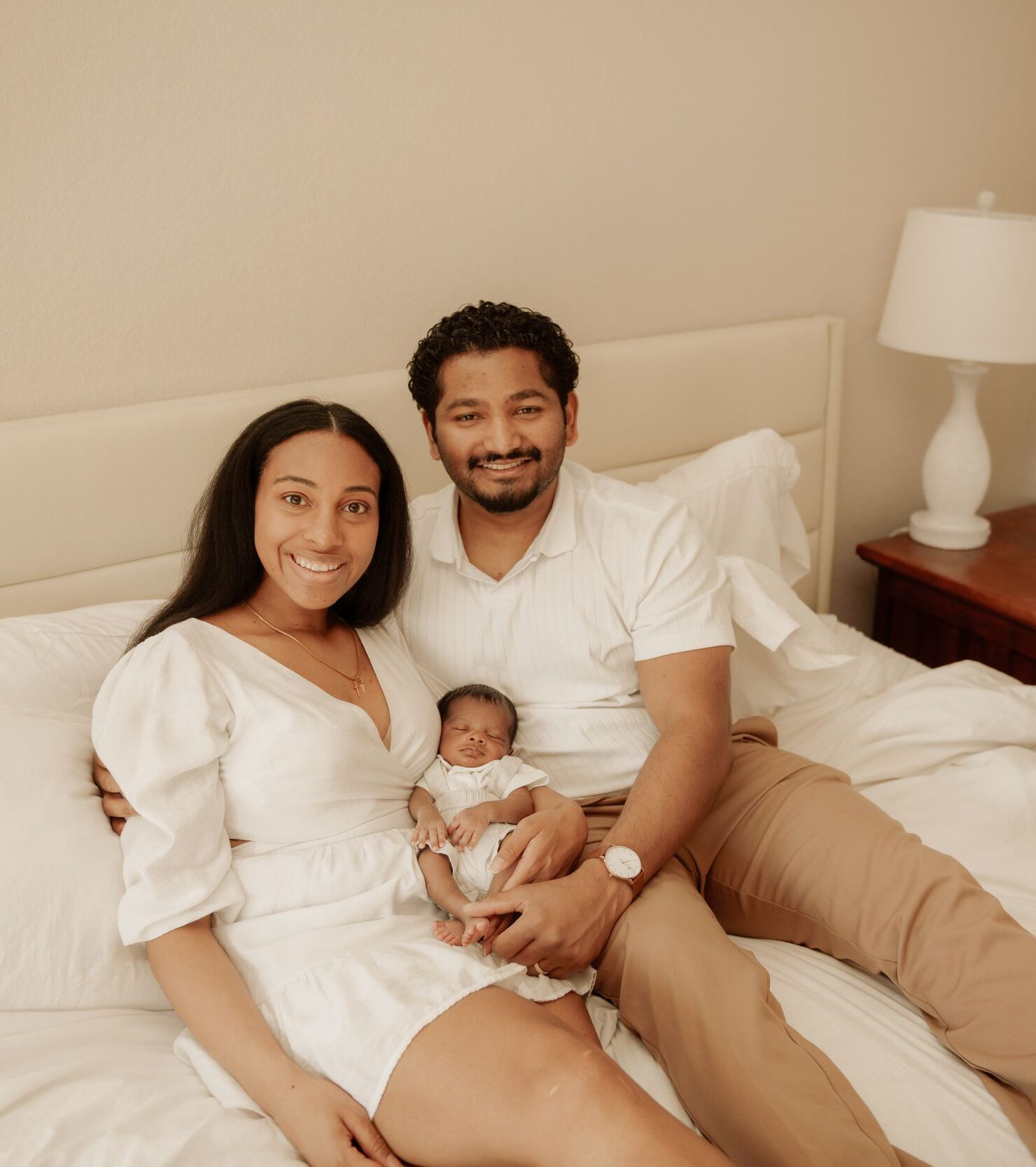 Parents sitting on a bed with their sleeping newborn baby.