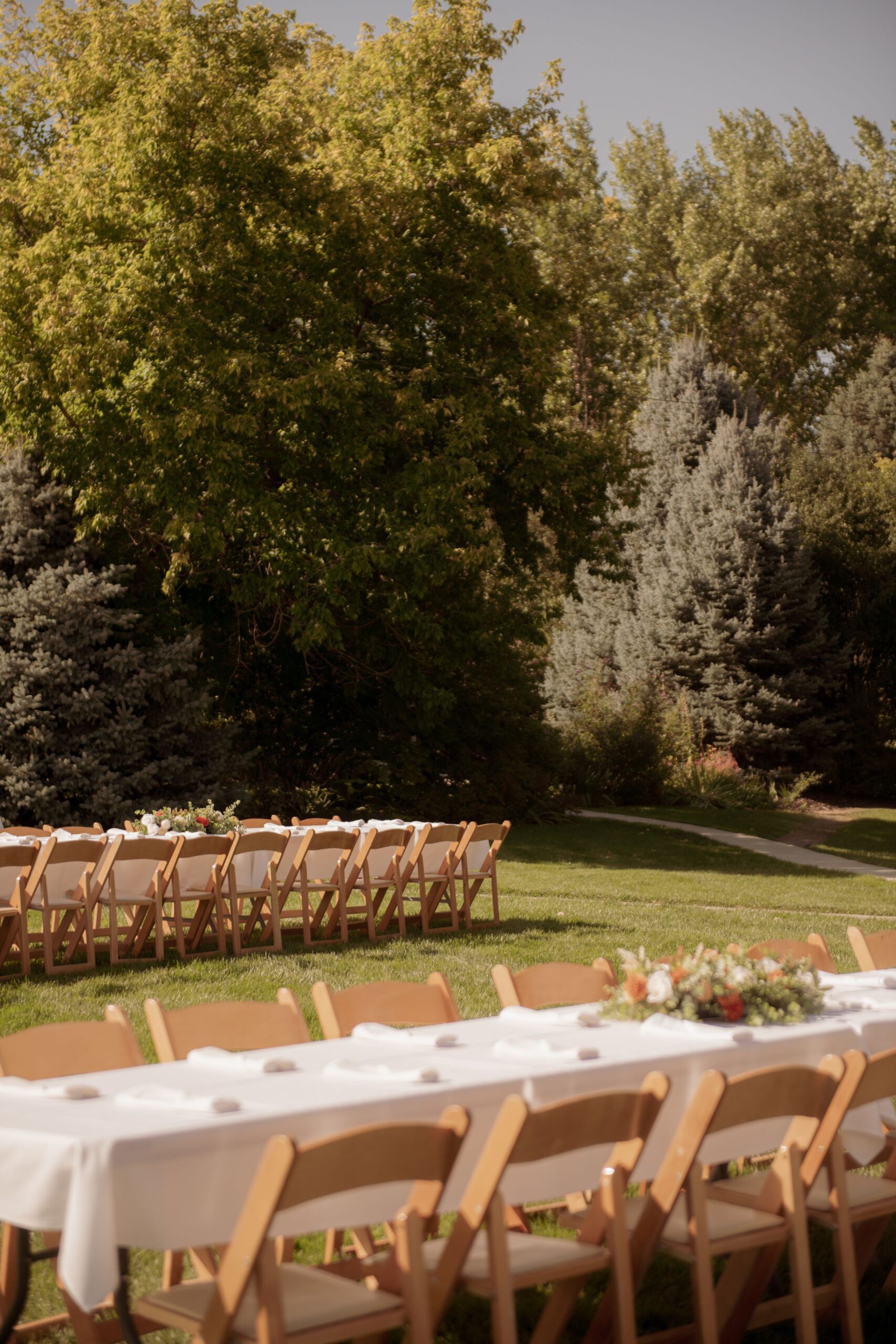 Outdoor wedding reception tables and wooden chairs set up on a lawn surrounded by trees.