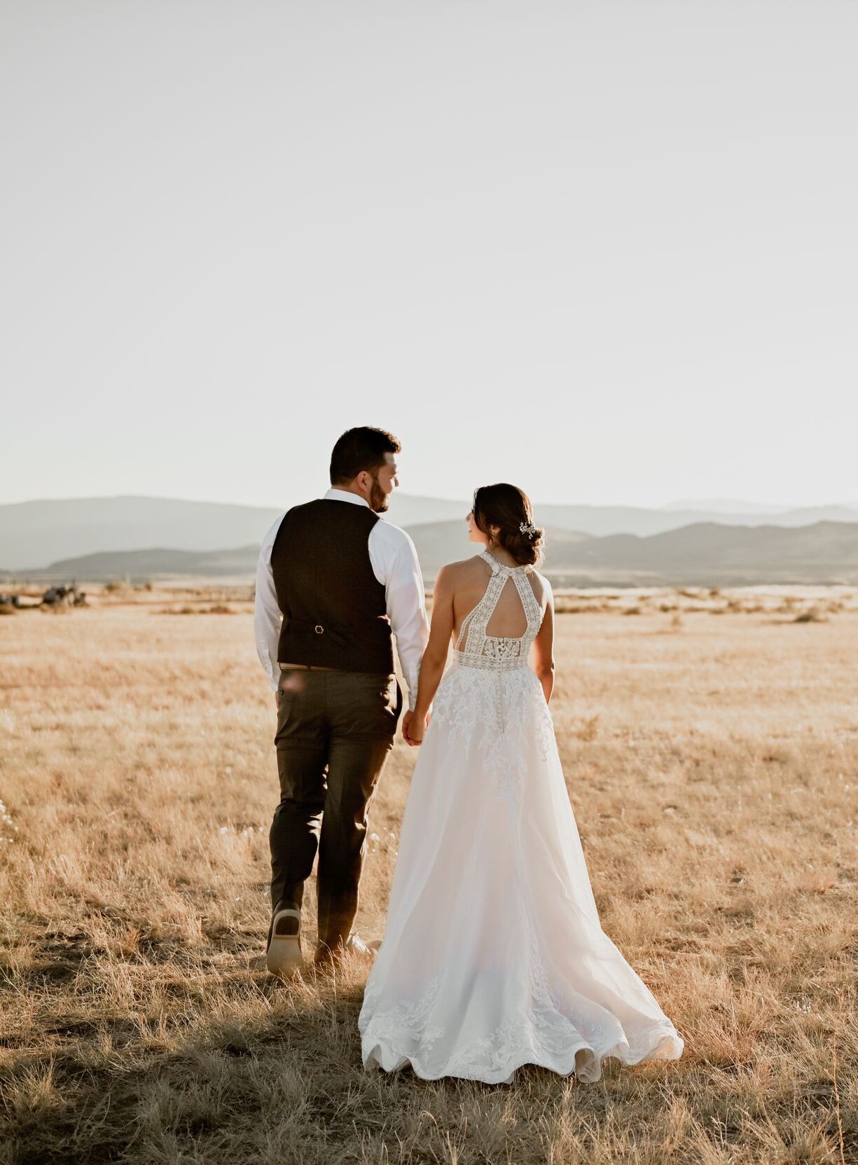 Bride and groom walking through a golden field at sunset, seen from behind