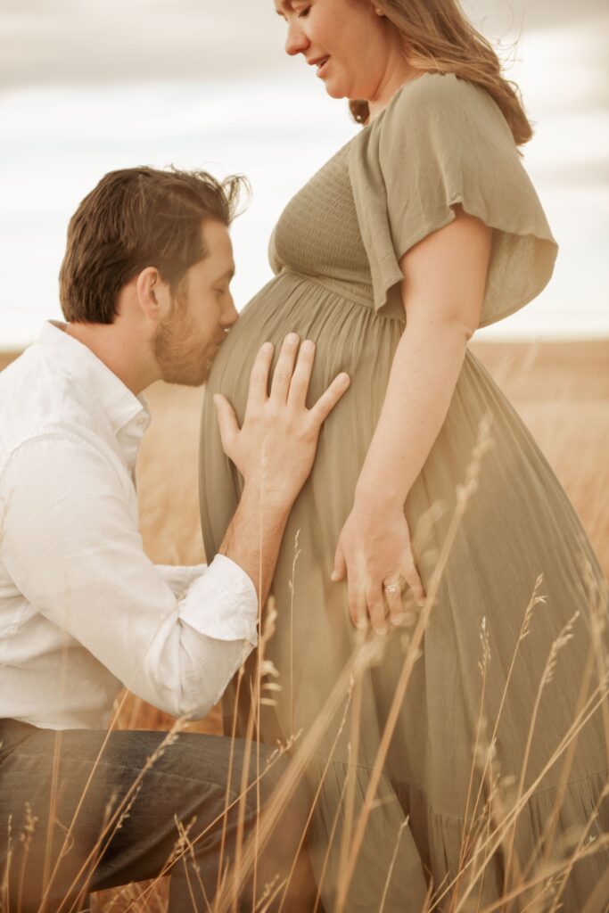 Expectant father kissing his partner's baby bump in a grassy field.