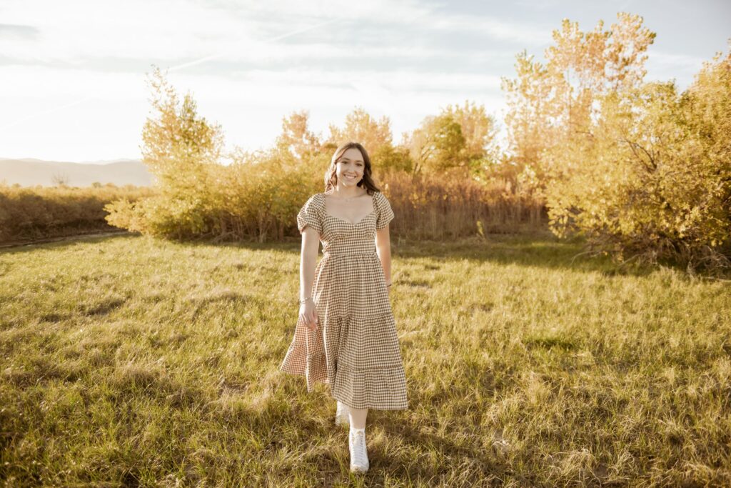 A woman in a plaid dress smiling in a golden autumn field at sunset.
