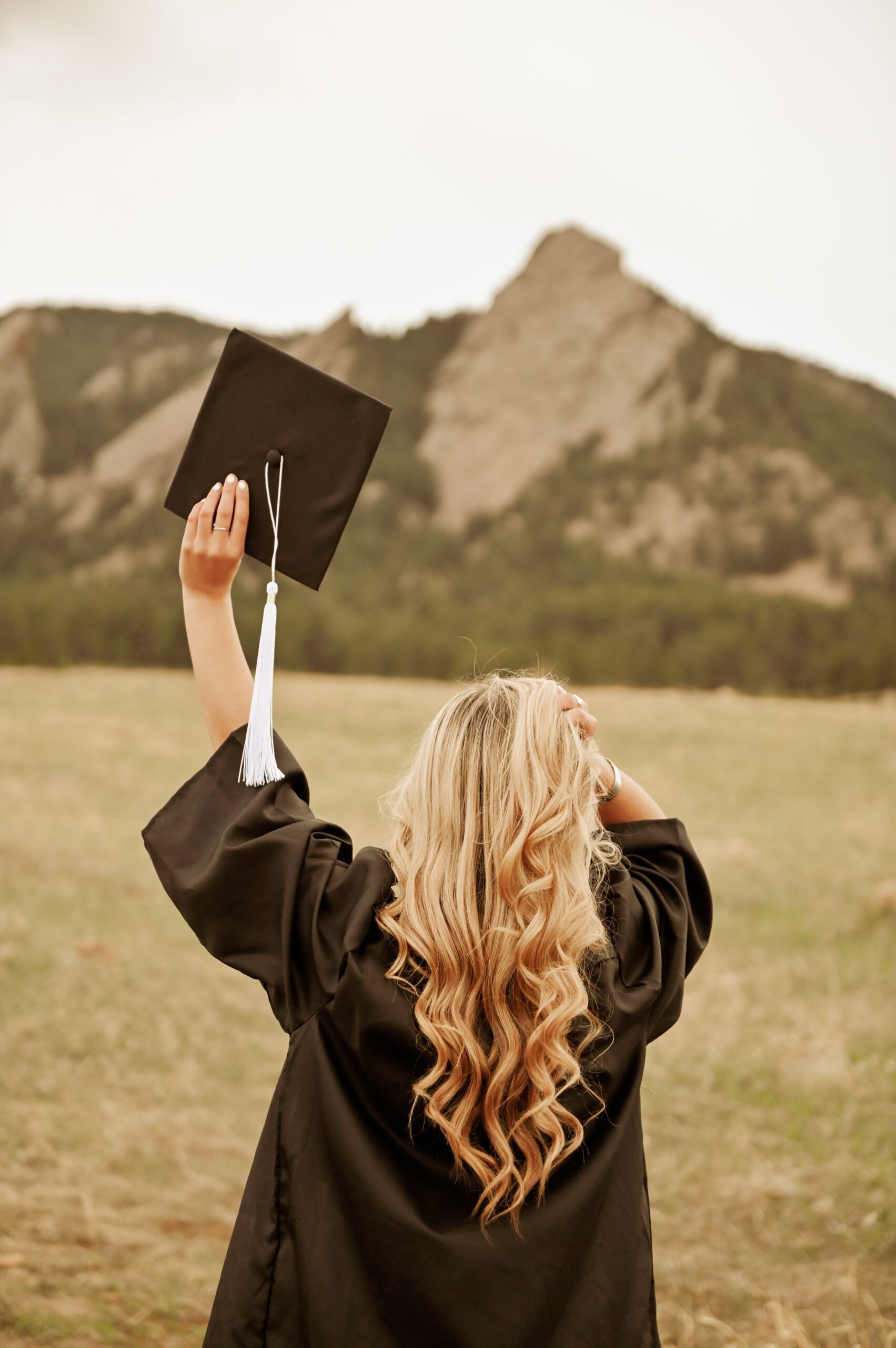 Graduate holding up her cap in a mountain field.