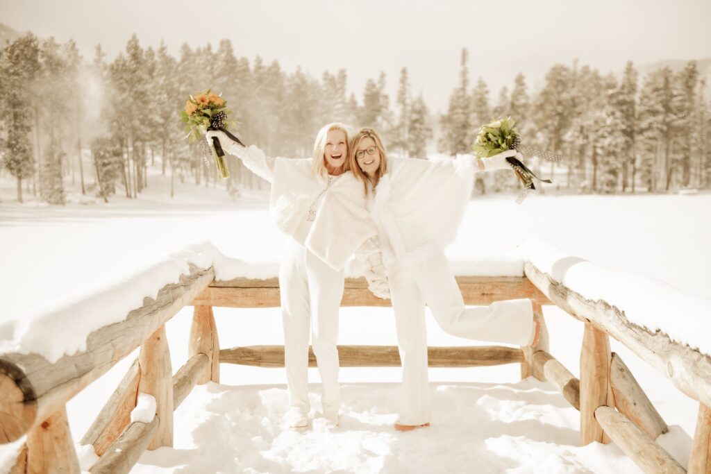 Two brides in matching white winter outfits laughing together in the snow, holding colorful bouquets.