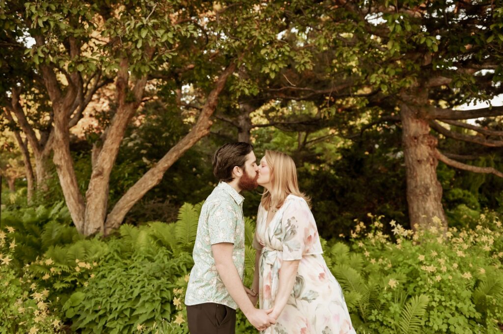 Couple kissing while holding hands in a lush green garden.
