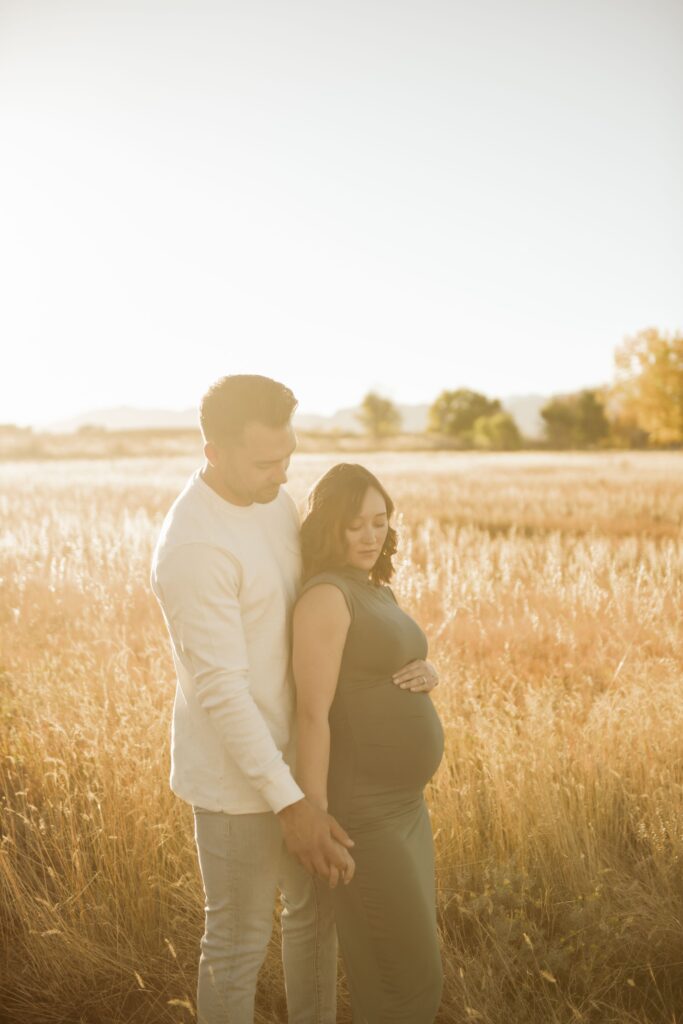 Couple standing together in a sunlit field during a maternity photoshoot.