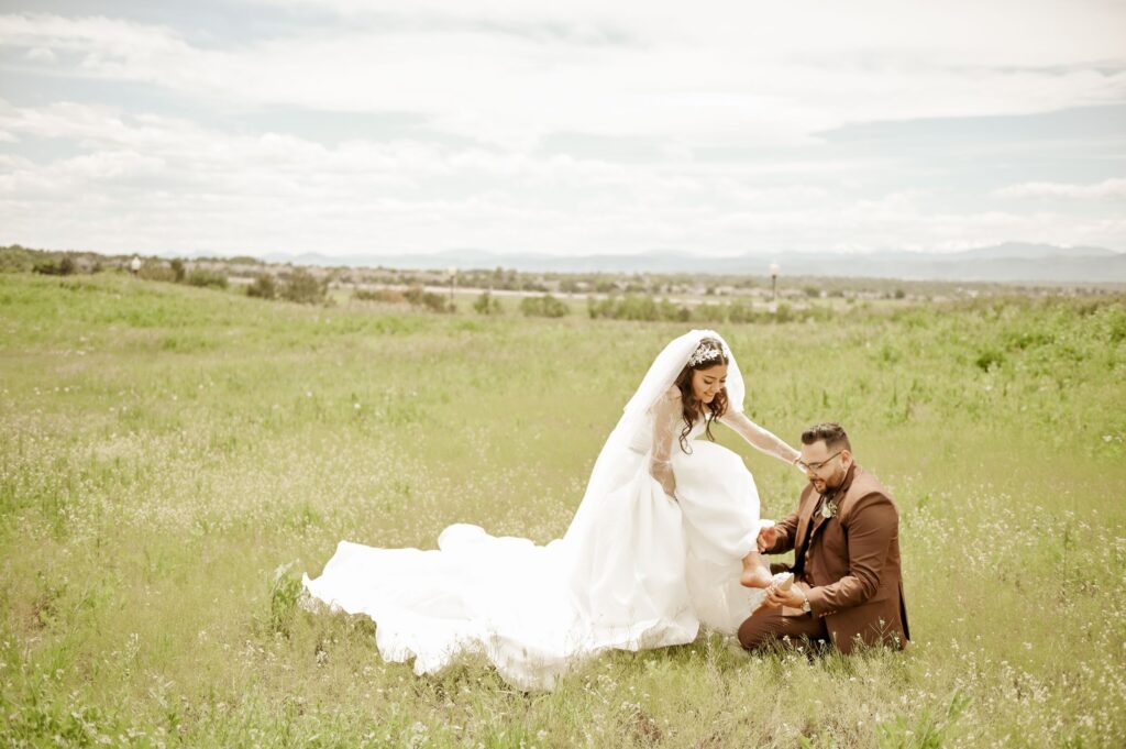 Groom helping the bride with her shoe in a grassy field during their wedding photos.