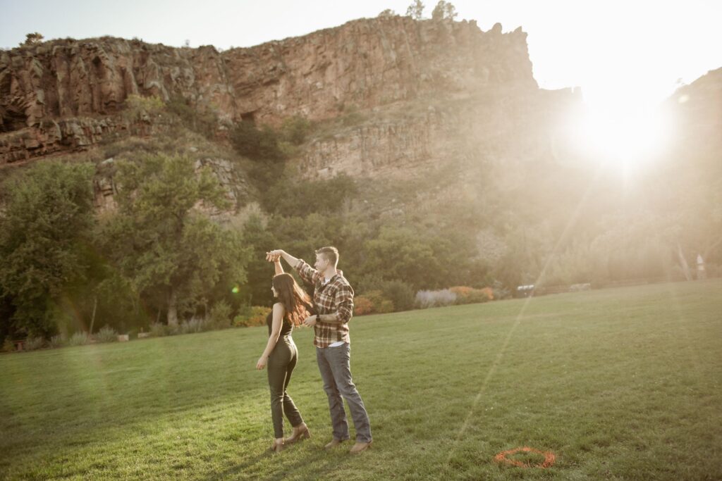 Man twirling his partner while dancing in a sunlit field by cliffs.
