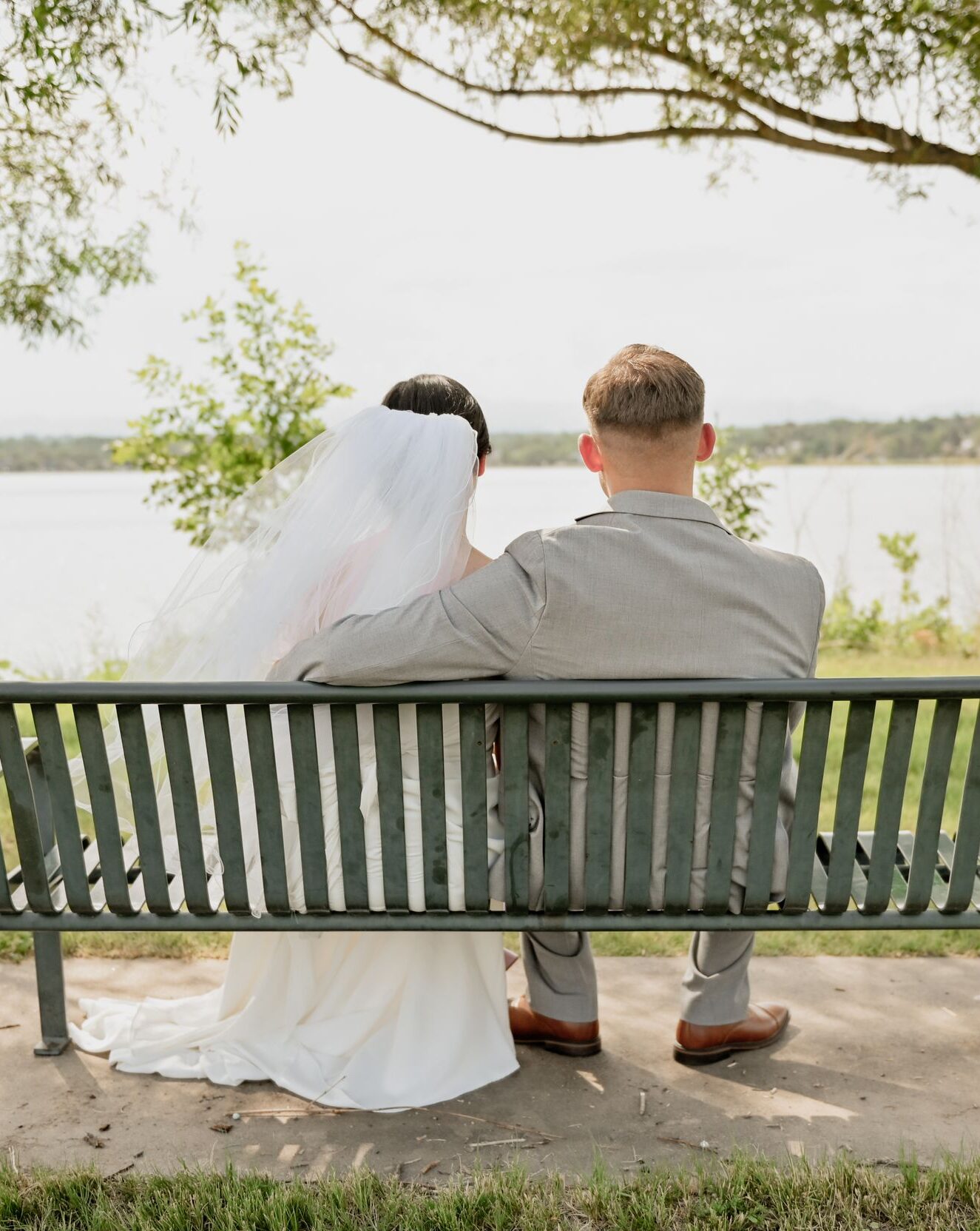 Bride and groom seen from behind, sitting close together on a park bench facing a lake.
