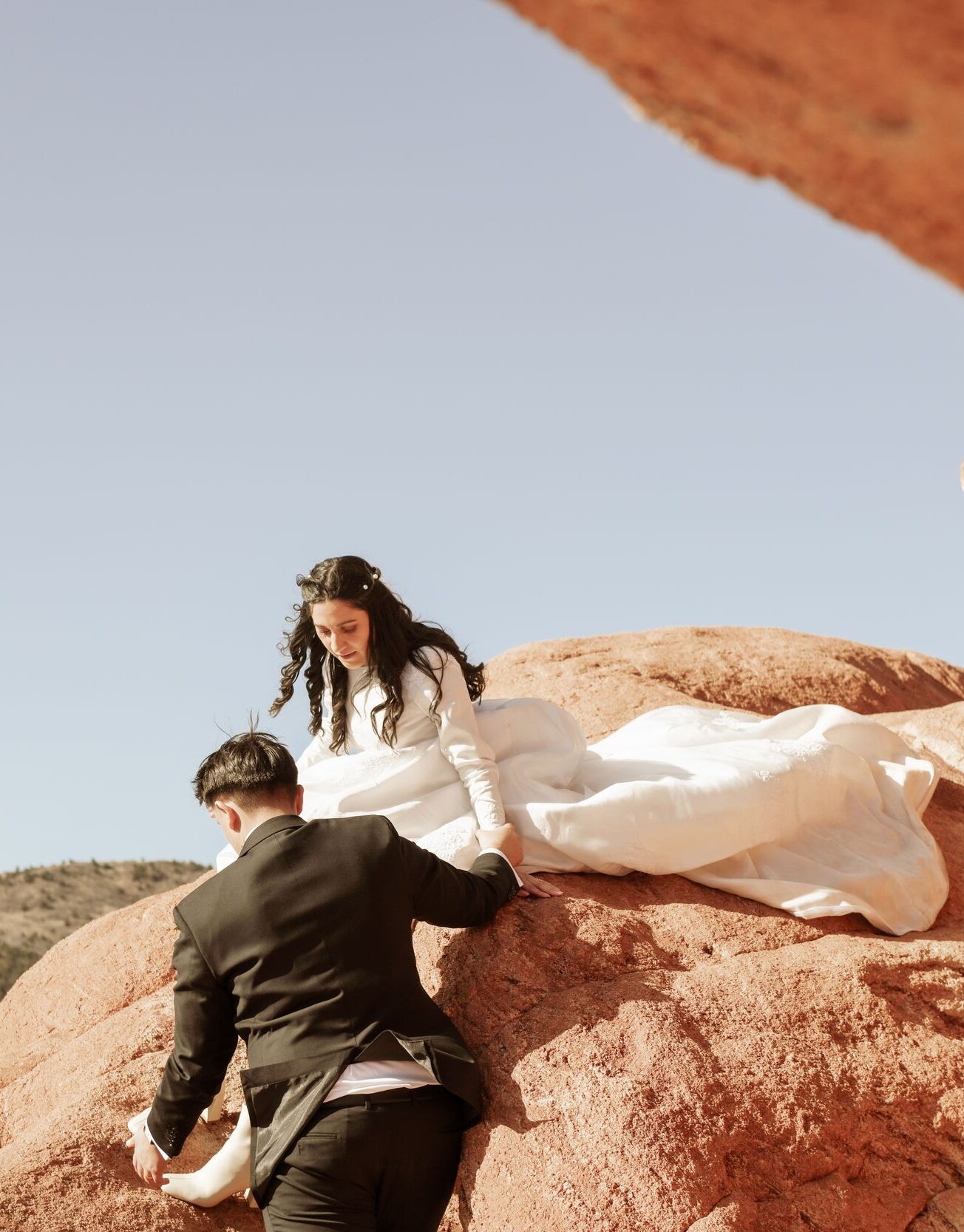 Groom helping bride climb down a large red rock formation.