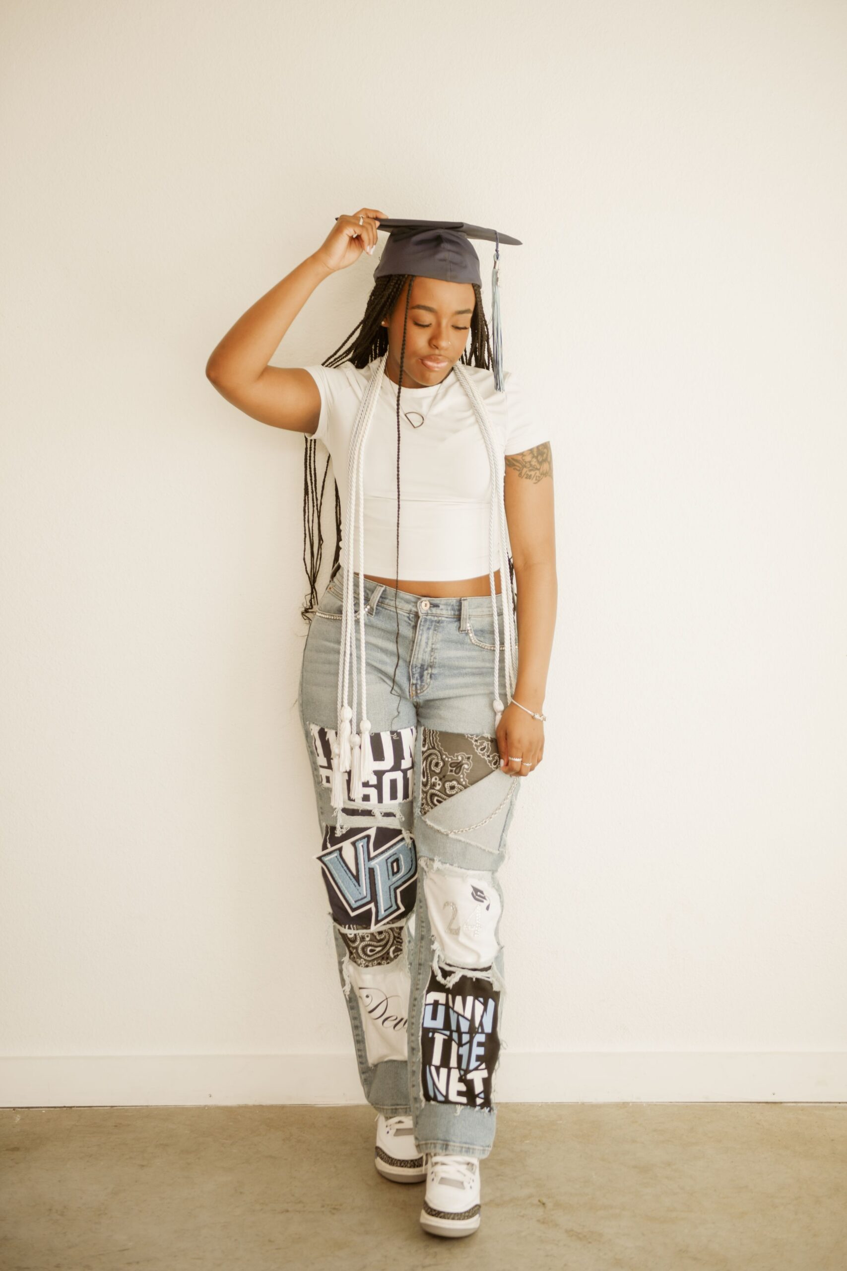 Graduate wearing a cap and honor cords posing in a white top and patchwork jeans against a plain wall.