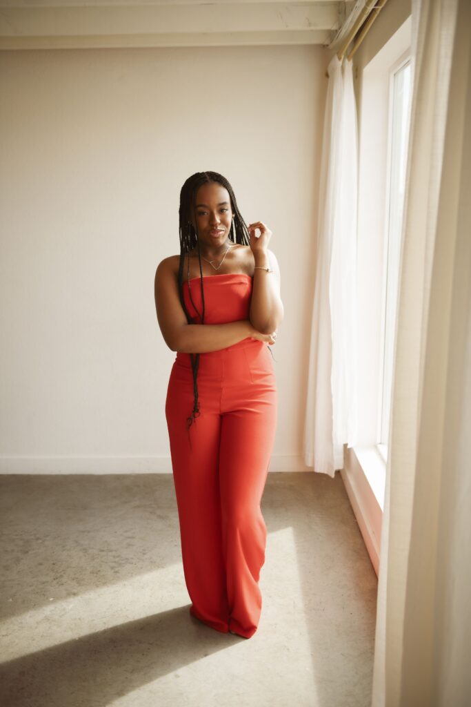 Woman in a red jumpsuit posing by a sunlit studio window.