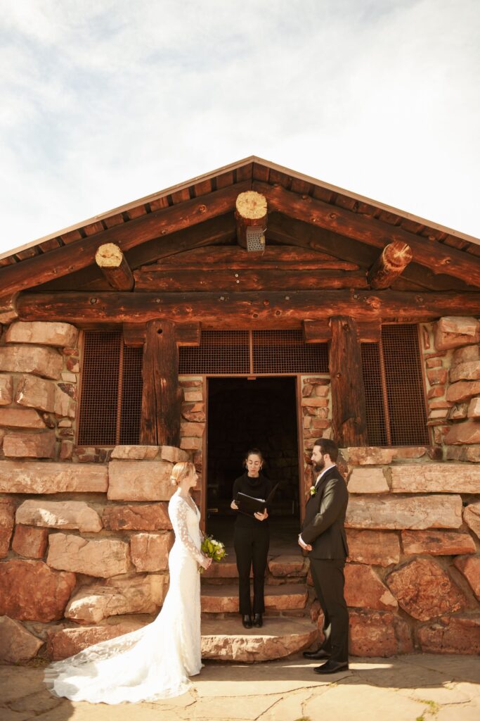 Bride and groom exchanging vows with an officiant outside a stone cabin.