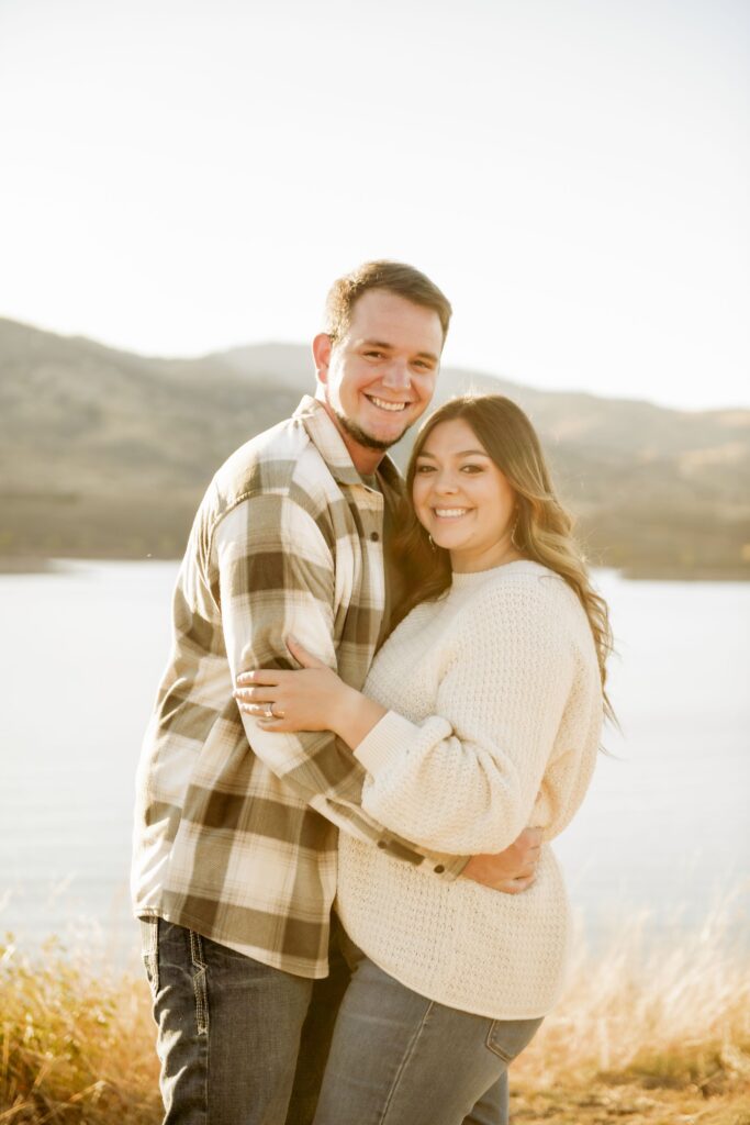Couple smiling and embracing by a lake at sunset.