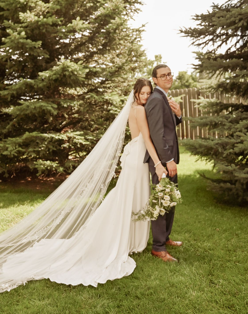 Bride and groom posed back-to-back among evergreen trees, her cathedral veil trailing on the grass.